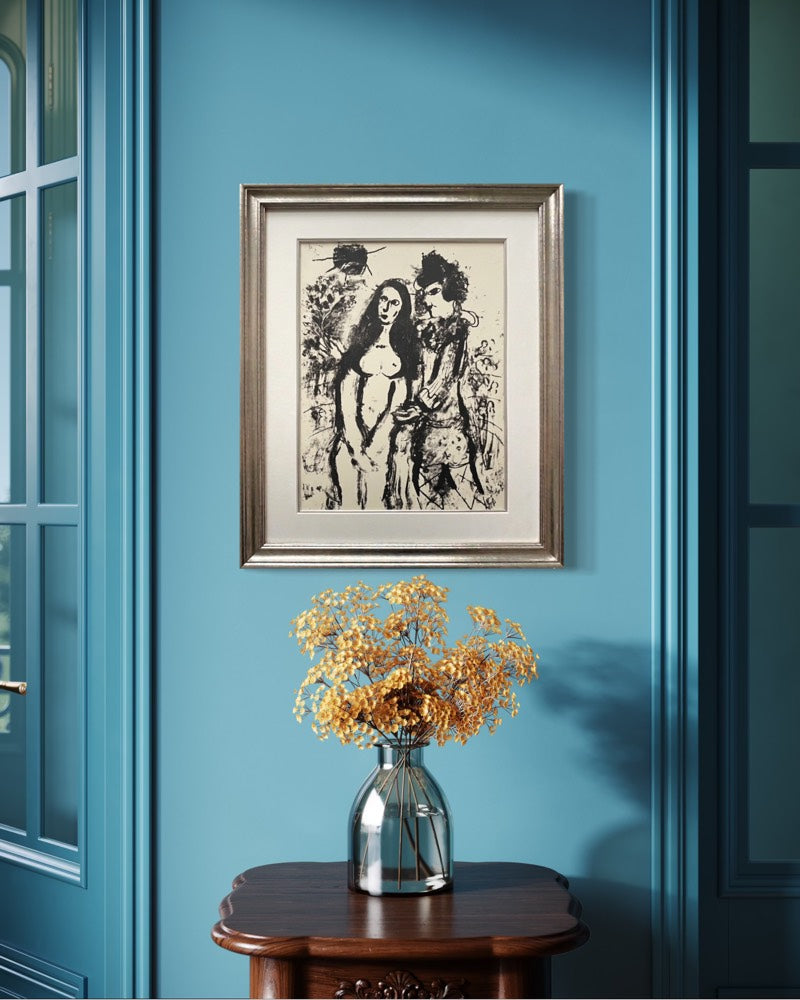 A framed black and white art print depicting two figures, styled on a blue wall above a wooden table with a vase of dried yellow flowers.