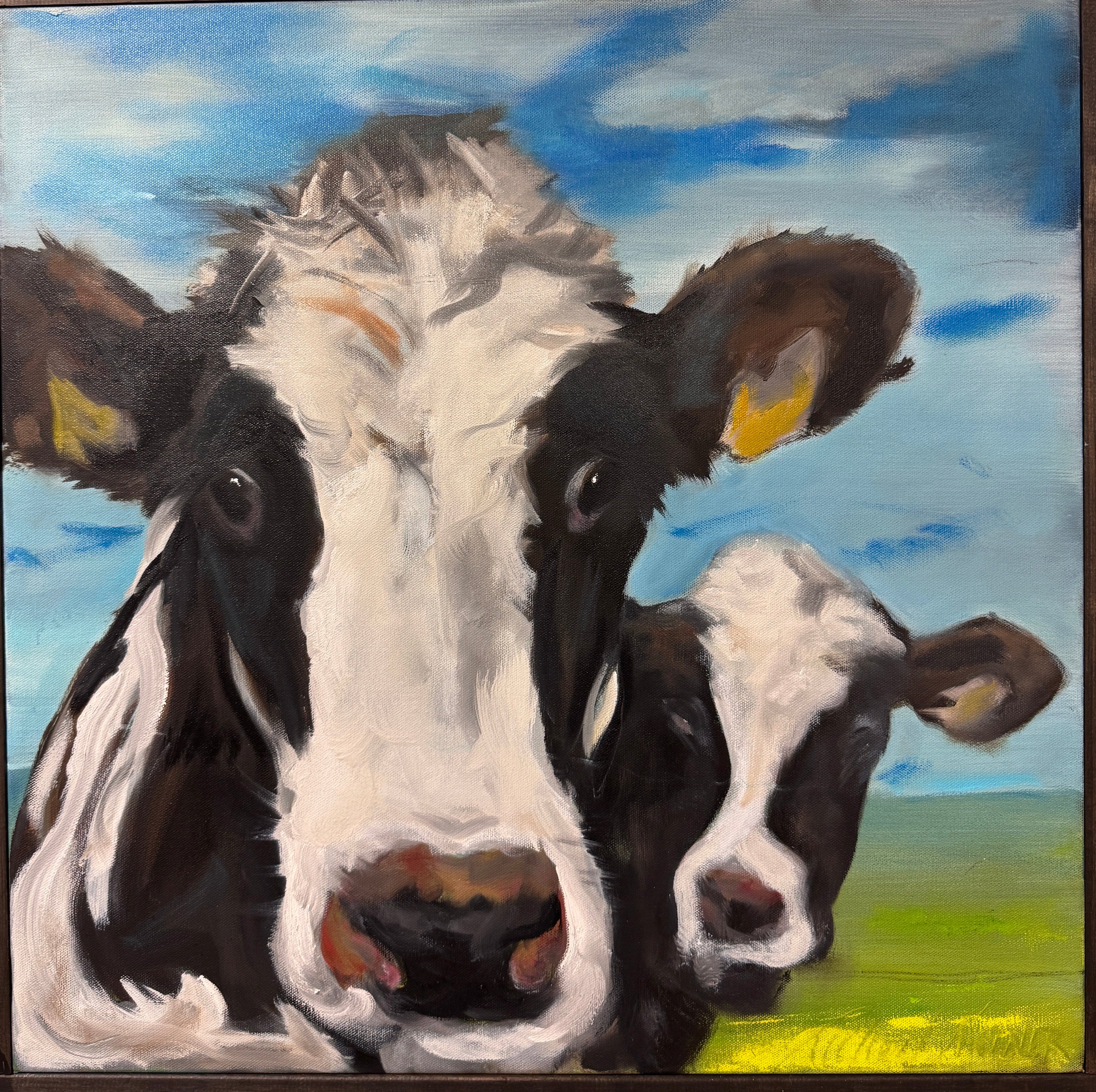 A square canvas painting of two black-and-white cows in a field under a blue sky, displayed on an easel in an art studio.
