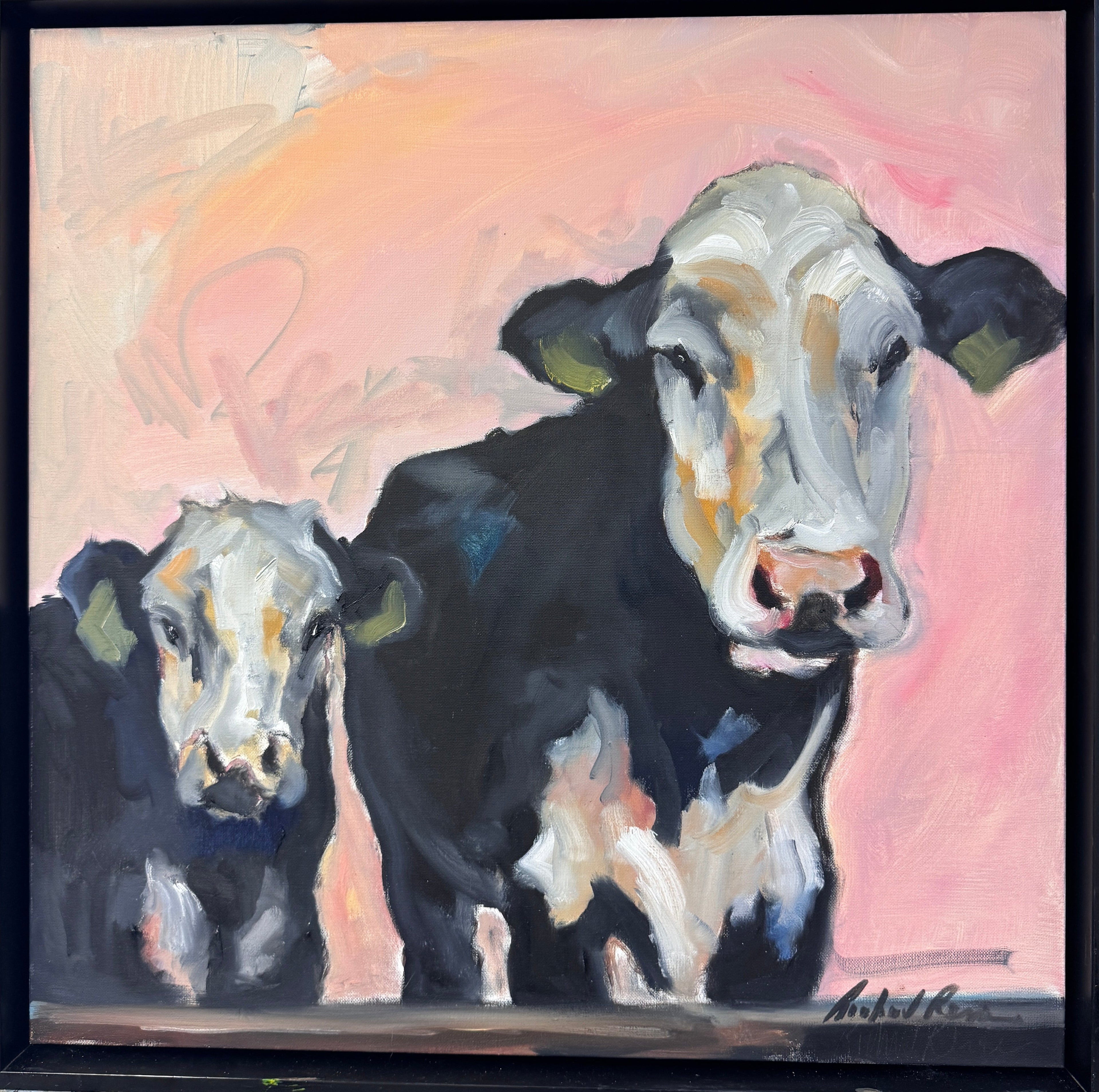 A framed canvas painting of two black and white cows against a pink and peach background, displayed on an easel in an art studio.